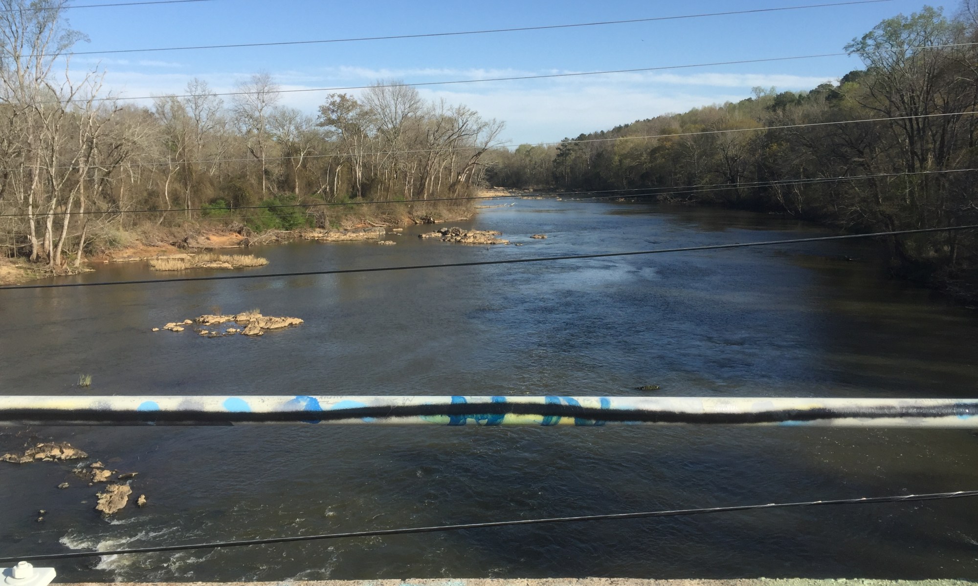 Overlooking the Haw River from a concrete bridge on a clear day. Graffiti on the bridge reads “THE RIVER KNOWS”