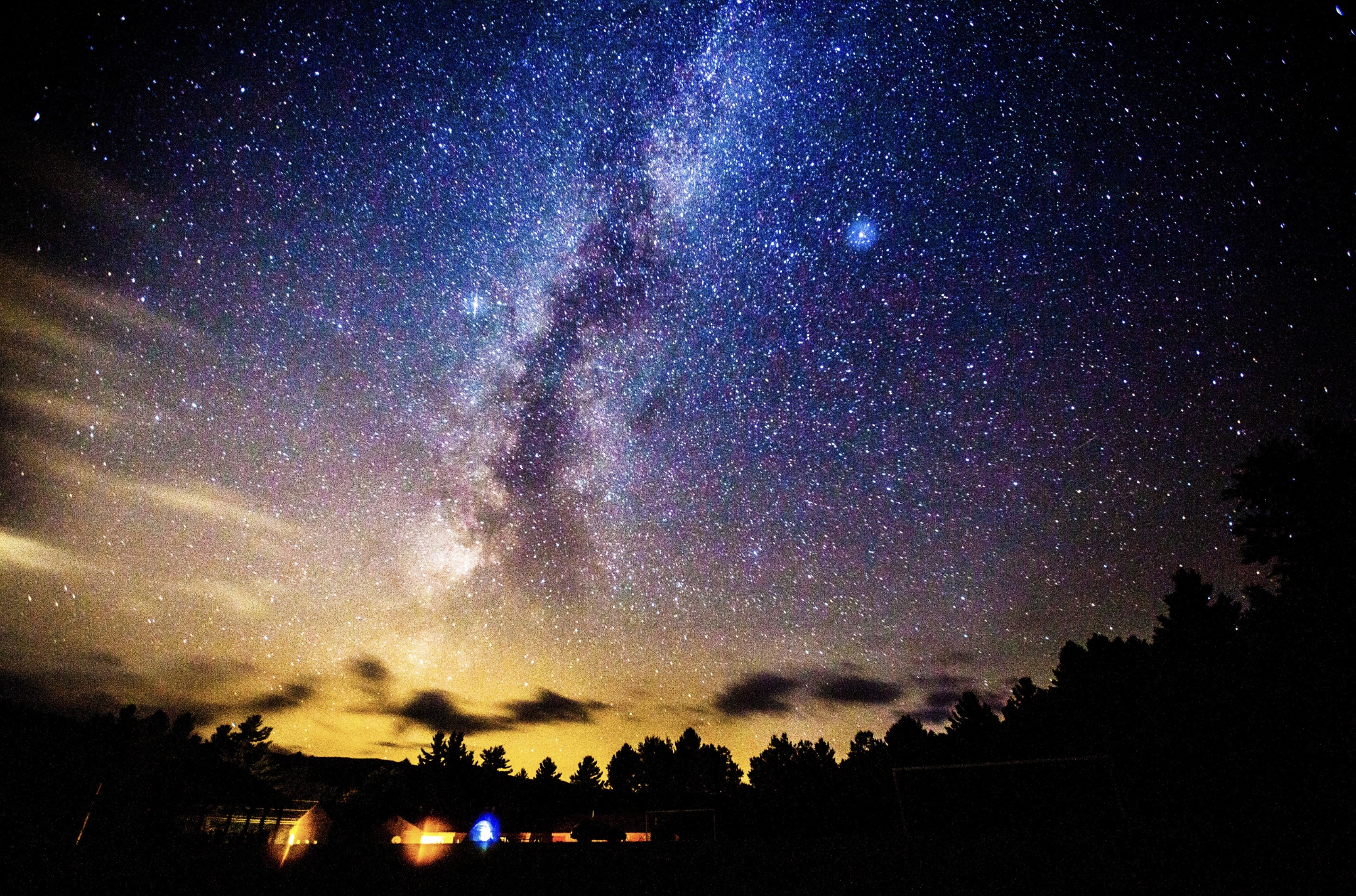 a long exposure photograph taken at night of a big sky where you can see the milky way clearly and millions of stars. The sky looks almost blue and there is some yellow entering in at the horizon. 