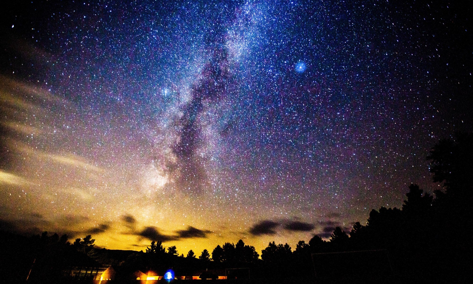 a long exposure photograph taken at night of a big sky where you can see the milky way clearly and millions of stars. The sky looks almost blue and there is some yellow entering in at the horizon.
