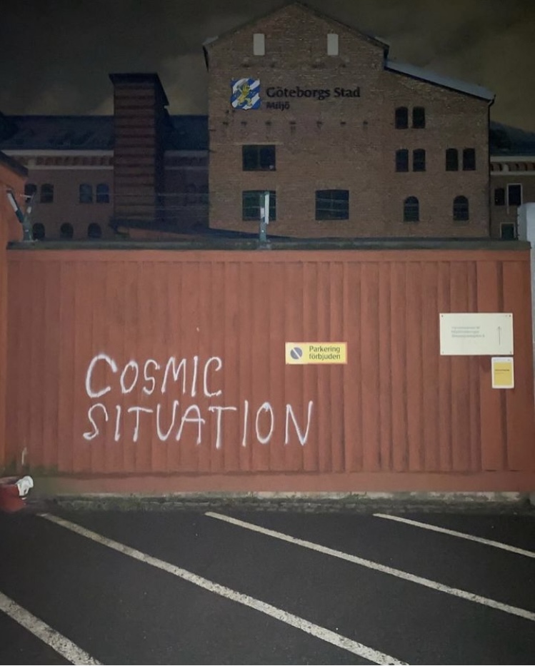 photo taken at night in a parking lot of a brown wall with white graffiti across that says “COSMIC SITUATION”