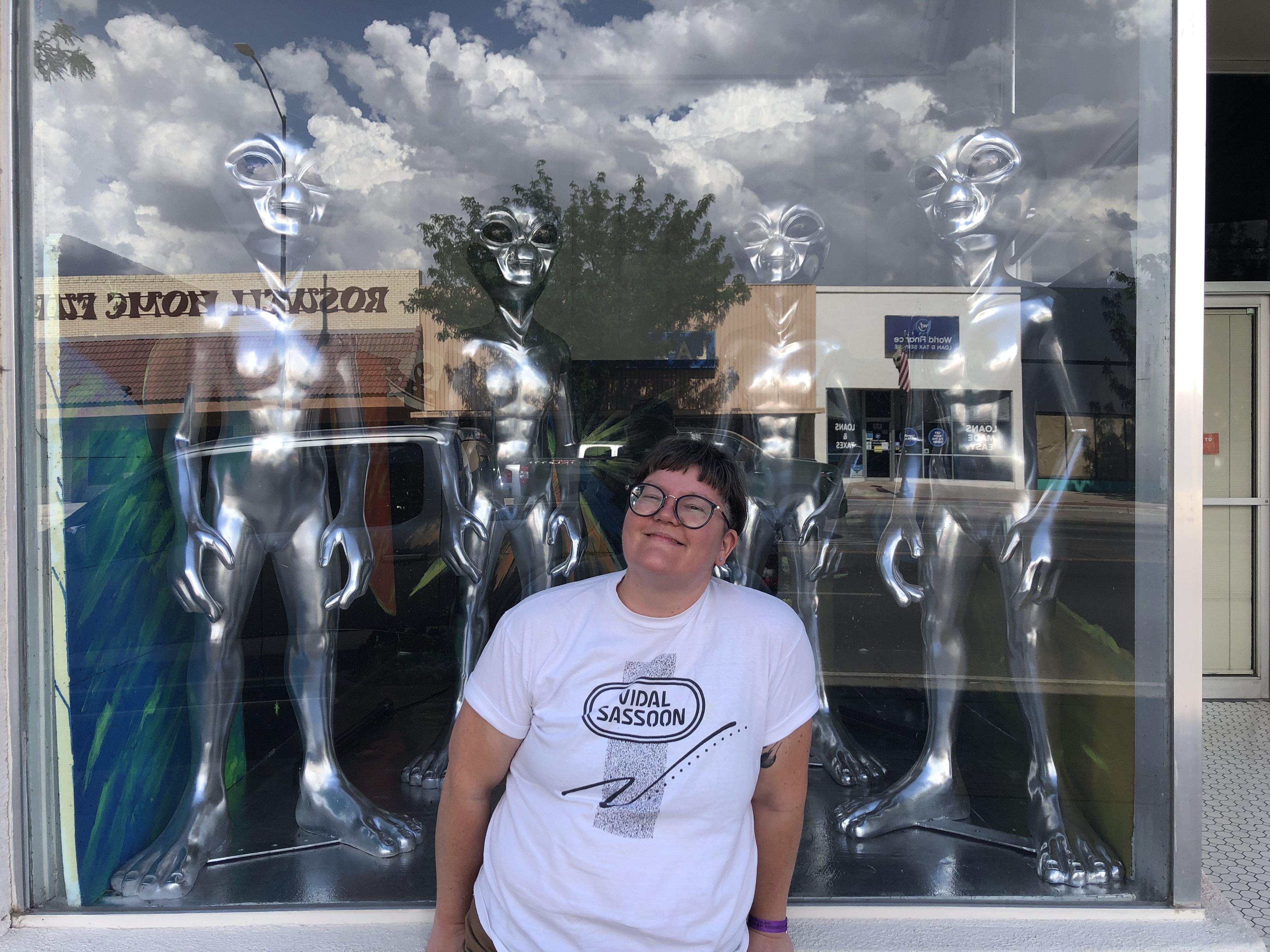 Photograph of Evan smiling big in front of a storefront window in Roswell, NM with 4 life size silver alien statues looking over them. Clouds are reflecting nicely in the glass. 