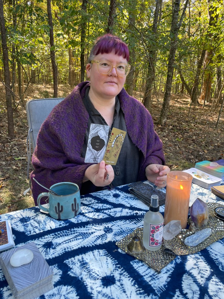 Photograph of Evan holding up two tarot cards while sitting outside at a table covered in indigo blue dyed fabric with a variety of tarot and oracle decks along with a candle and crystals. 