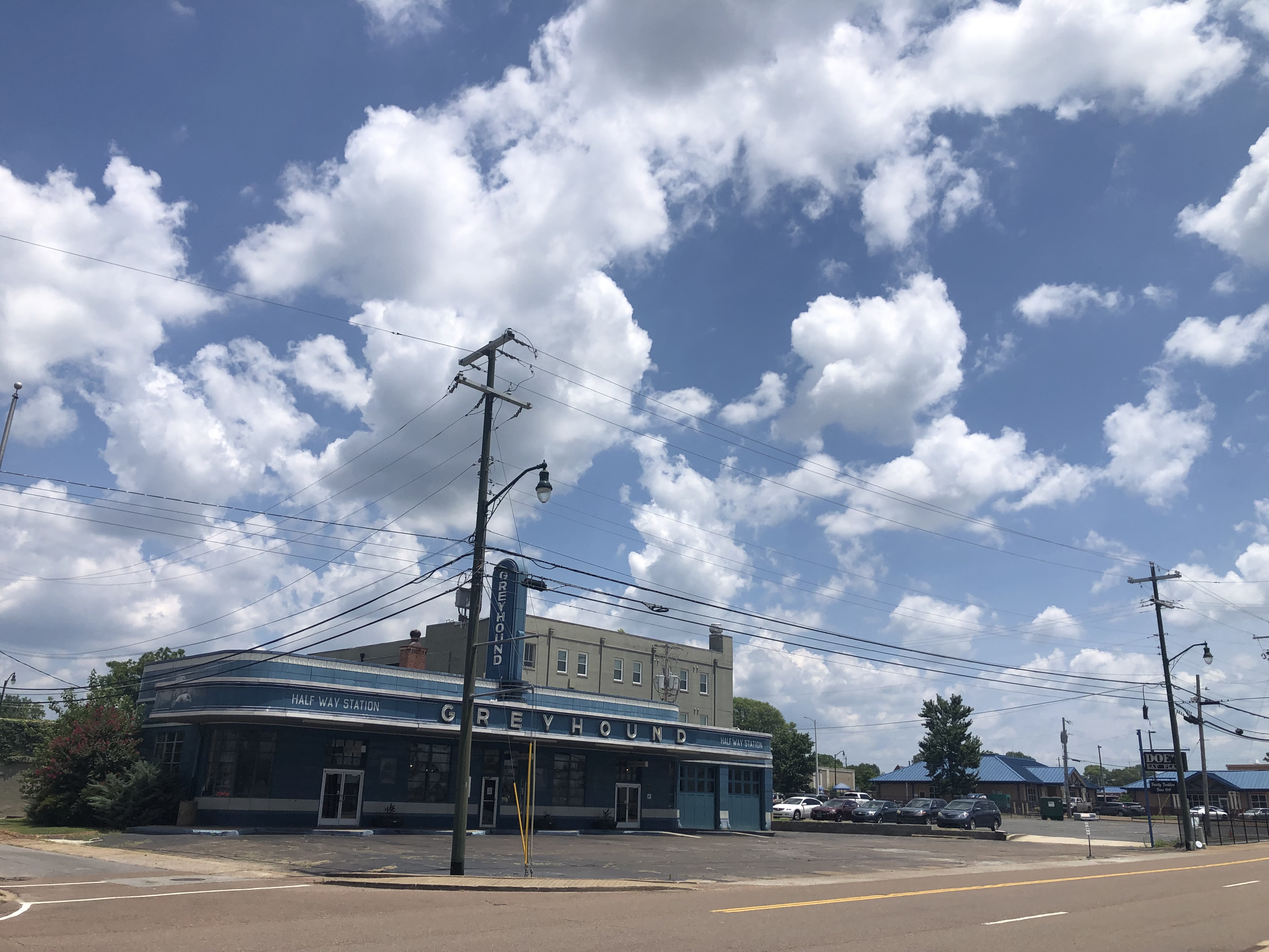 Photograph of an old Greyhound bus station in TN that looks like it is from the 1940’s. The sky is a vibrant blue with lots of white puffy clouds above.